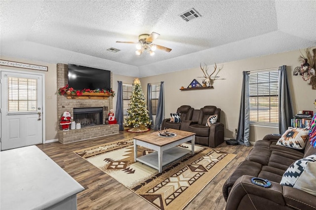 living room with a brick fireplace, ceiling fan, wood-type flooring, and a tray ceiling