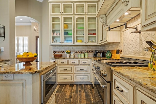 kitchen featuring cream cabinets, dark wood-style floors, arched walkways, appliances with stainless steel finishes, and glass insert cabinets