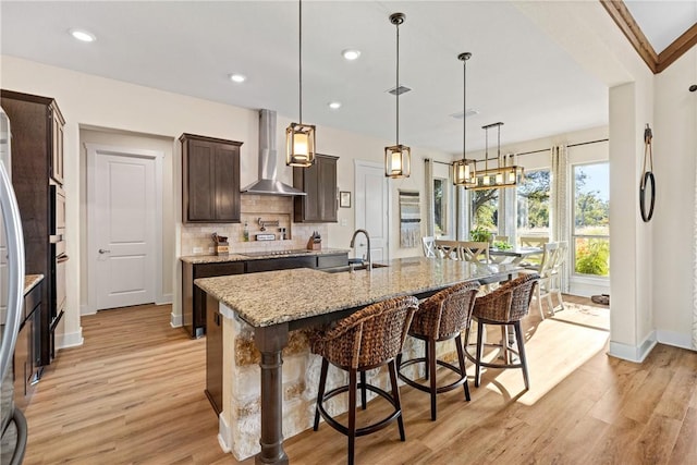 kitchen featuring pendant lighting, sink, a kitchen island with sink, a kitchen breakfast bar, and wall chimney exhaust hood