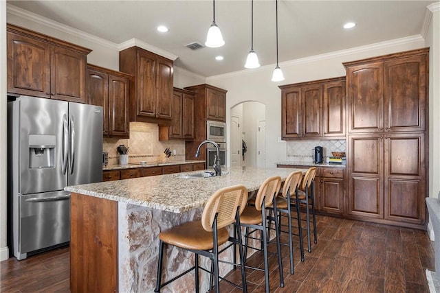 kitchen with a kitchen island with sink, arched walkways, a sink, stainless steel appliances, and dark wood-type flooring