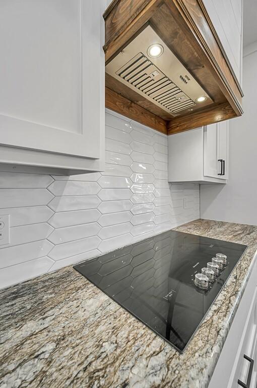 kitchen with decorative backsplash, light stone countertops, premium range hood, black electric cooktop, and white cabinets