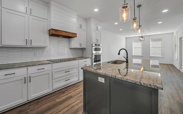 kitchen with black electric stovetop, dark wood-type flooring, sink, a center island with sink, and hanging light fixtures