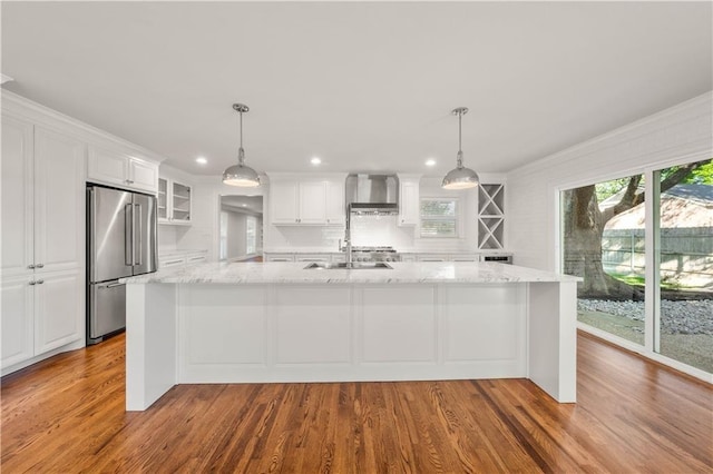 kitchen with high end fridge, white cabinets, hanging light fixtures, and wall chimney range hood