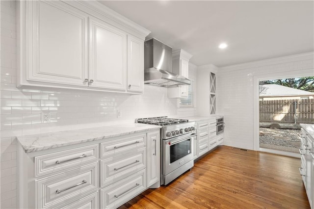 kitchen with decorative backsplash, stainless steel appliances, wall chimney range hood, white cabinets, and light hardwood / wood-style floors