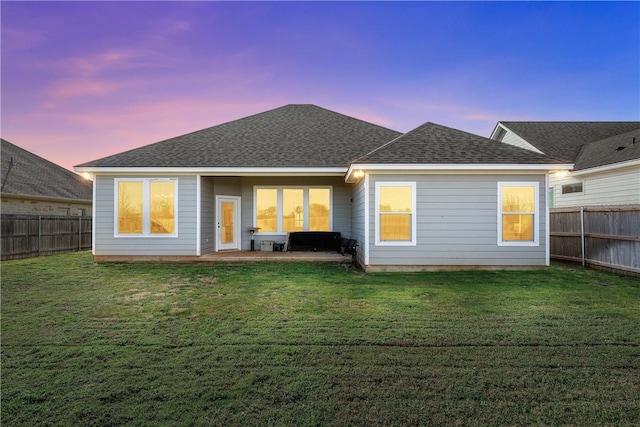 back of house at dusk with a lawn, a fenced backyard, and a shingled roof