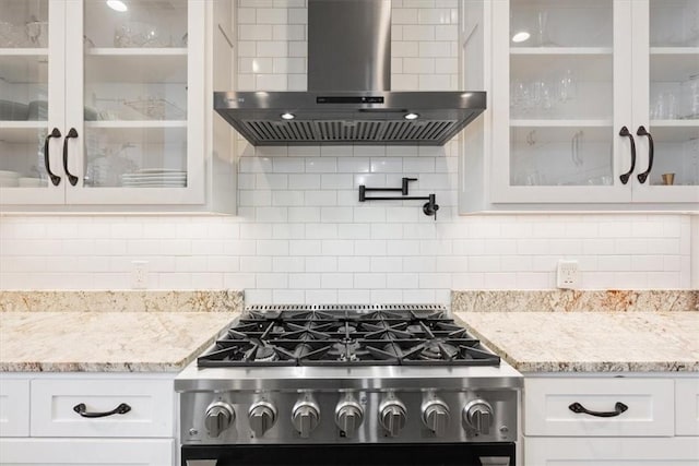 kitchen featuring glass insert cabinets, white cabinets, wall chimney range hood, light stone countertops, and range