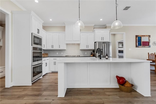 kitchen featuring appliances with stainless steel finishes, hardwood / wood-style flooring, white cabinets, hanging light fixtures, and an island with sink