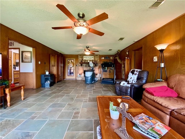 living room featuring ceiling fan, wood walls, a wood stove, and a textured ceiling