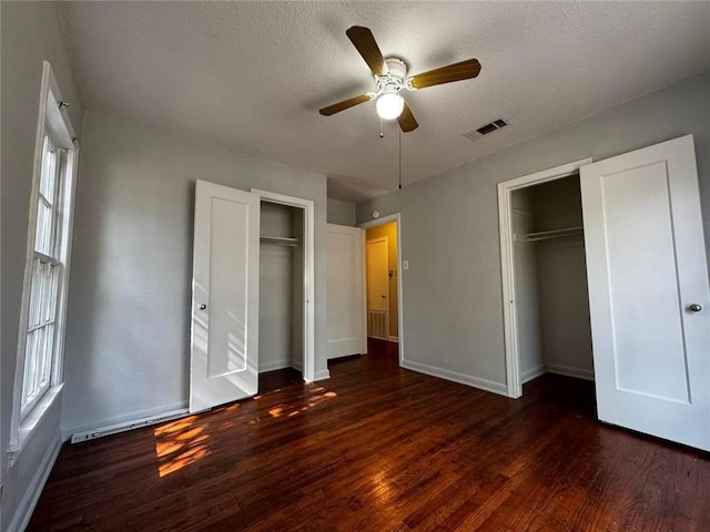 unfurnished bedroom with a textured ceiling, ceiling fan, dark wood-type flooring, and multiple closets