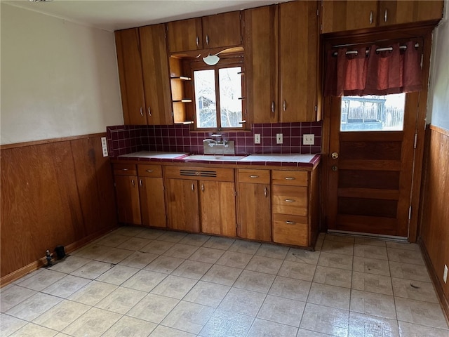 kitchen with a sink, tile countertops, wooden walls, brown cabinetry, and wainscoting