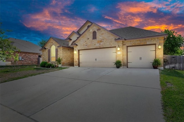 french provincial home with roof with shingles, concrete driveway, an attached garage, fence, and stone siding