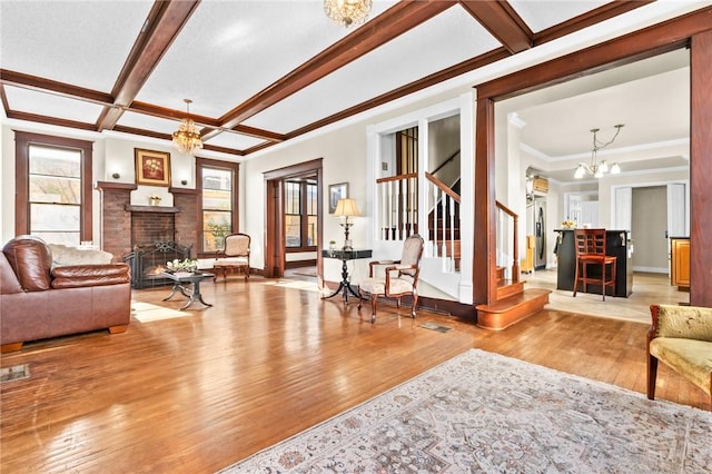 living area featuring coffered ceiling, stairway, hardwood / wood-style floors, a brick fireplace, and a notable chandelier