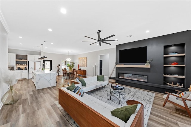 living room with built in shelves, light wood-type flooring, ceiling fan with notable chandelier, and ornamental molding