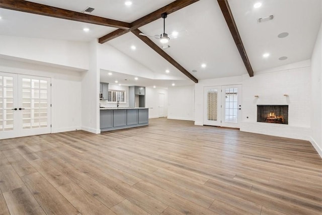 unfurnished living room featuring a brick fireplace, ceiling fan, light wood-type flooring, french doors, and beamed ceiling