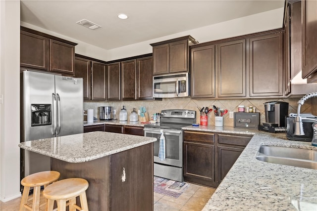 kitchen featuring dark brown cabinetry, light stone countertops, stainless steel appliances, a kitchen bar, and decorative backsplash