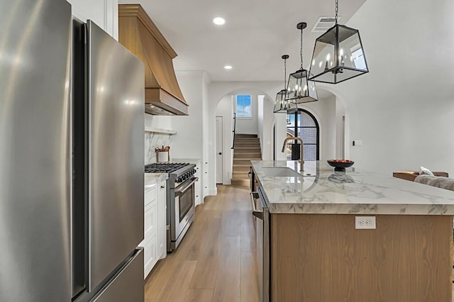 kitchen with light stone counters, stainless steel appliances, light hardwood / wood-style flooring, hanging light fixtures, and an island with sink