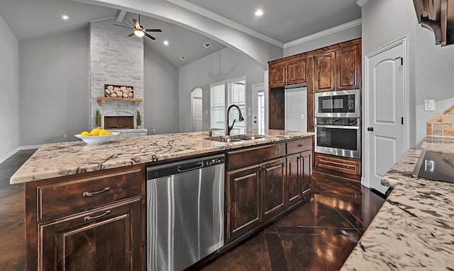 kitchen featuring a stone fireplace, vaulted ceiling, an island with sink, appliances with stainless steel finishes, and dark brown cabinetry