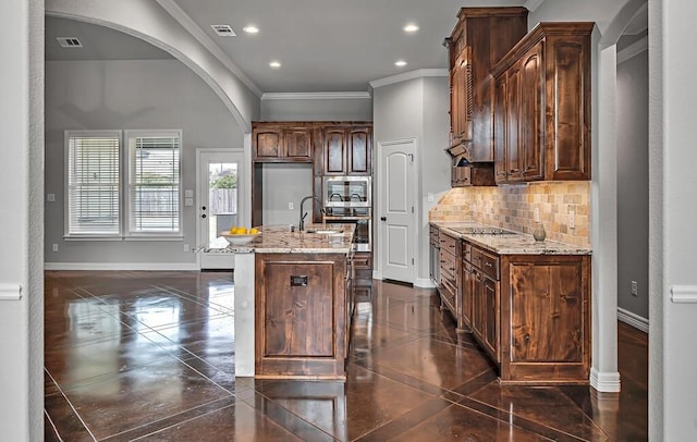 kitchen with sink, tasteful backsplash, light stone counters, an island with sink, and ornamental molding