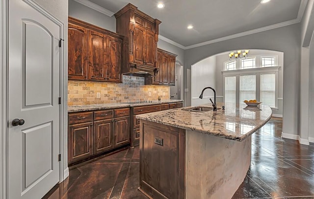 kitchen featuring light stone counters, dark brown cabinets, crown molding, sink, and a center island with sink