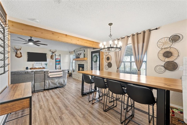 dining space with a textured ceiling, light hardwood / wood-style floors, ceiling fan with notable chandelier, and a stone fireplace