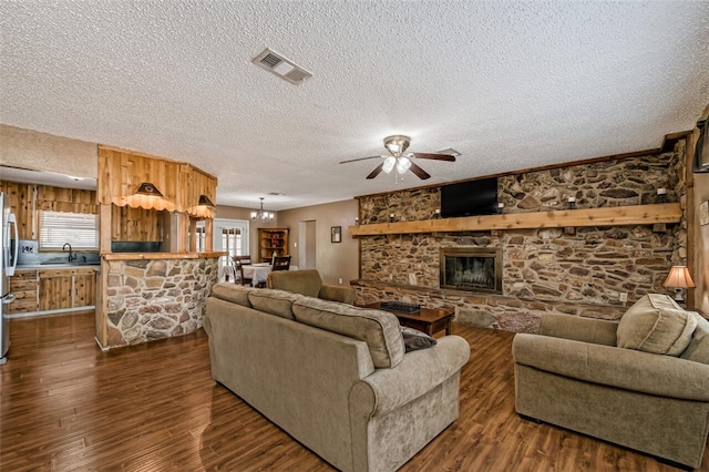 living room featuring sink, dark wood-type flooring, a stone fireplace, a textured ceiling, and ceiling fan with notable chandelier