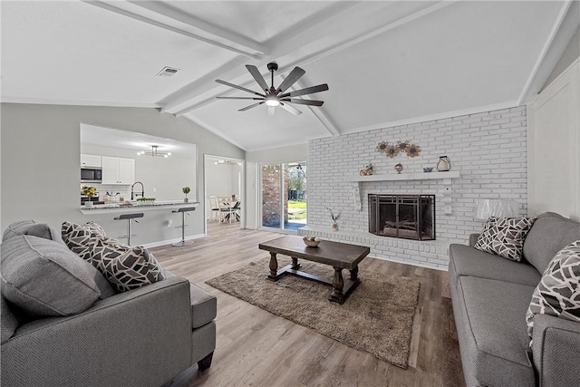 living room featuring vaulted ceiling with beams, light wood-type flooring, a brick fireplace, and ceiling fan