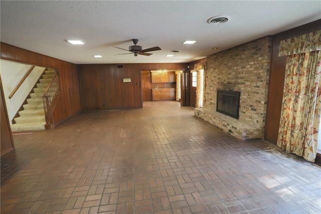 unfurnished living room featuring ceiling fan, a textured ceiling, wooden walls, and a brick fireplace