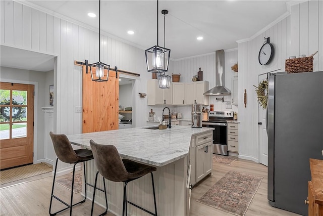 kitchen featuring wall chimney range hood, sink, a barn door, an island with sink, and appliances with stainless steel finishes