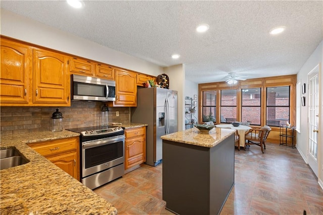 kitchen with ceiling fan, stainless steel appliances, a center island, light stone counters, and decorative backsplash