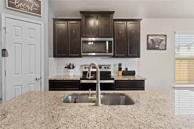 kitchen with dark brown cabinetry, backsplash, light stone countertops, stainless steel appliances, and a sink