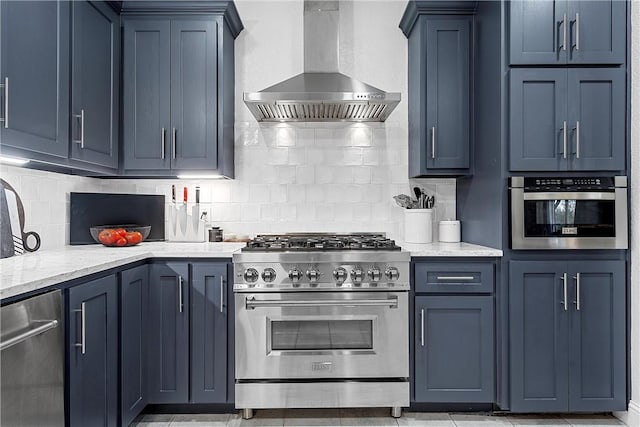 kitchen with blue cabinetry, stainless steel appliances, wall chimney range hood, light stone counters, and backsplash