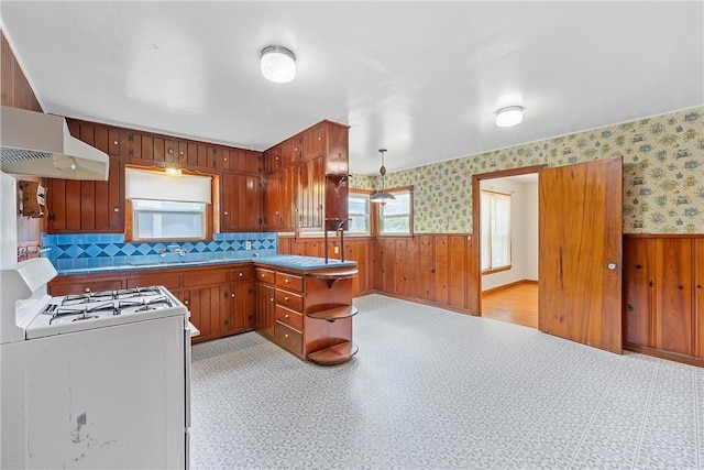 kitchen with kitchen peninsula, wooden walls, hanging light fixtures, white gas stove, and range hood