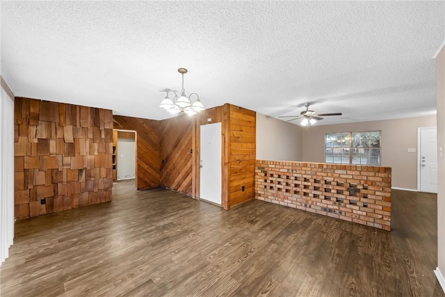 unfurnished living room with a textured ceiling, dark wood-type flooring, wooden walls, and ceiling fan with notable chandelier