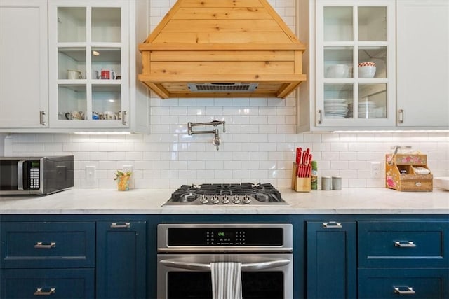 kitchen featuring blue cabinets, premium range hood, decorative backsplash, white cabinets, and appliances with stainless steel finishes