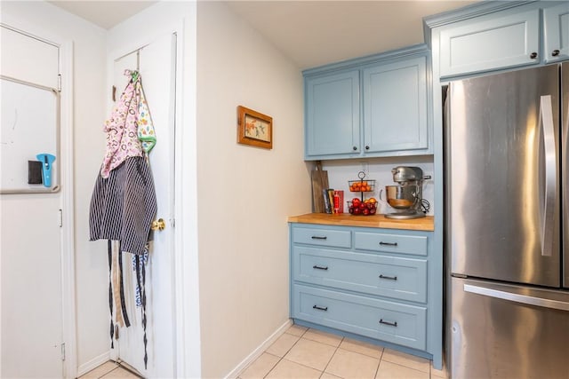 kitchen featuring blue cabinetry, butcher block counters, freestanding refrigerator, light tile patterned flooring, and baseboards