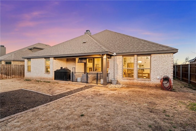 back of house at dusk featuring a fenced backyard, a chimney, a shingled roof, and brick siding