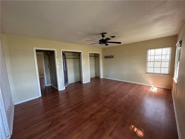 unfurnished bedroom with dark wood-type flooring, baseboards, multiple closets, and a ceiling fan