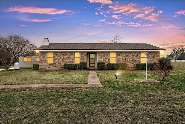 ranch-style home with a shingled roof, brick siding, a yard, and a chimney