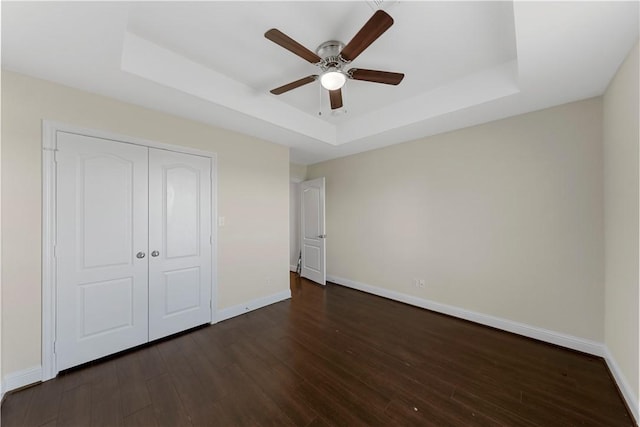 unfurnished bedroom with a tray ceiling, ceiling fan, a closet, and dark wood-type flooring