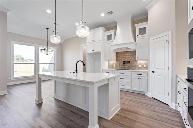 kitchen featuring pendant lighting, hardwood / wood-style floors, premium range hood, white cabinets, and an island with sink