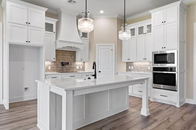 kitchen with appliances with stainless steel finishes, custom range hood, decorative light fixtures, a center island with sink, and white cabinetry