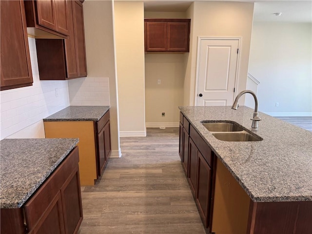 kitchen with a sink, baseboards, light wood finished floors, a center island with sink, and tasteful backsplash