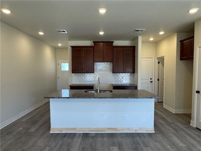 kitchen featuring dark wood-style flooring, a kitchen island with sink, a sink, dark stone counters, and baseboards