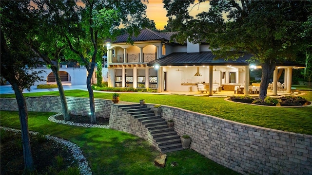 back house at dusk with an outdoor hangout area, a balcony, a patio, and a lawn