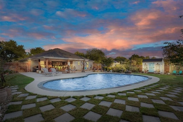 pool at dusk featuring a lawn, a patio area, and an outbuilding