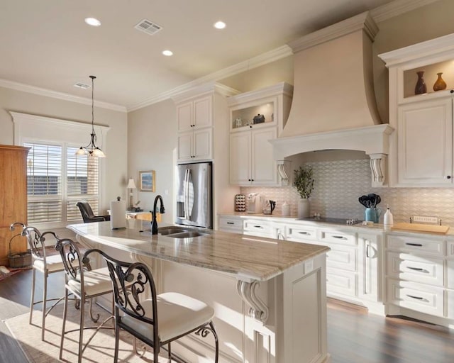 kitchen featuring sink, premium range hood, an island with sink, and stainless steel refrigerator with ice dispenser