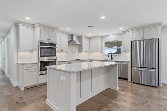 kitchen with light wood-type flooring, wall chimney exhaust hood, stainless steel appliances, white cabinetry, and a kitchen island