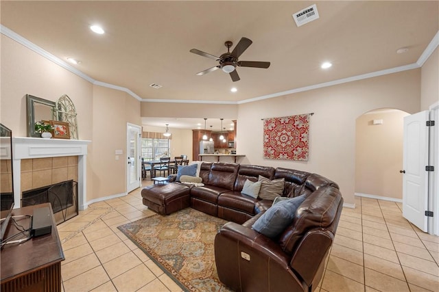 living room featuring a tiled fireplace, light tile patterned floors, crown molding, and ceiling fan