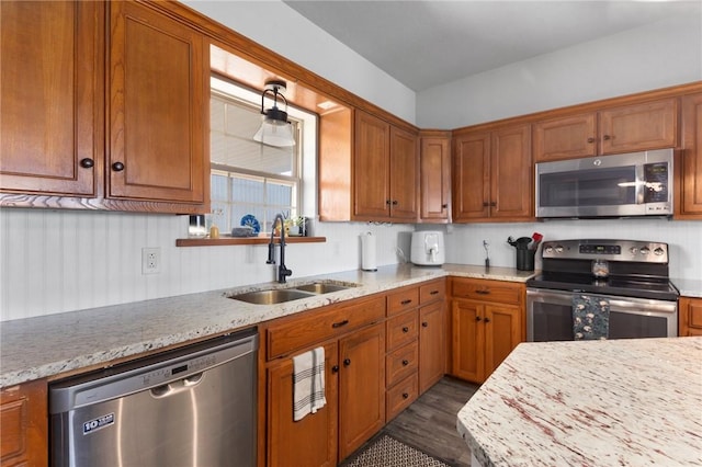 kitchen with brown cabinetry, appliances with stainless steel finishes, dark wood-style flooring, light stone countertops, and a sink