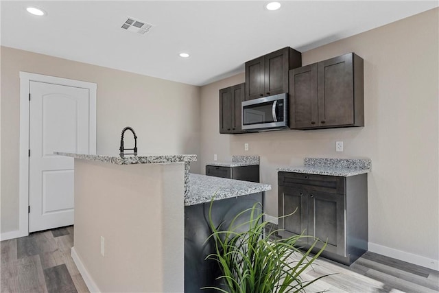 kitchen with light wood-type flooring, light stone counters, dark brown cabinetry, a kitchen island with sink, and sink